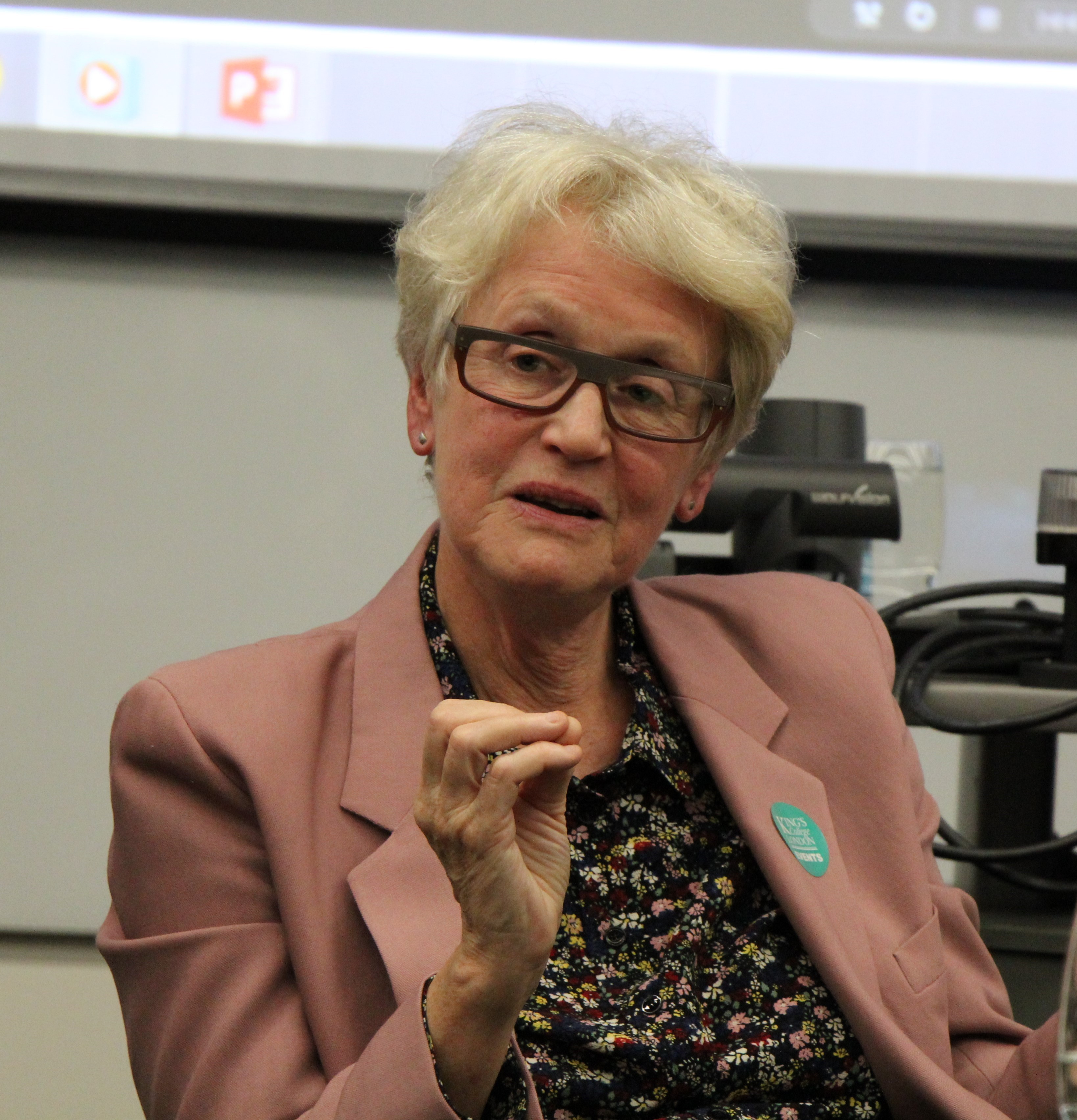 Color photograph of Barbara Taylor in a lecture hall wearing a pink suit jacket and glasses.