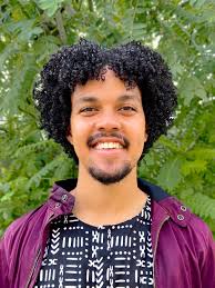Color headshot of Alonso Malik Pirio wearing a purple button down in front of greenery.